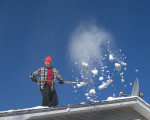 shoveller on roof with snow in the air against a blue sky